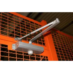 This bolt-on door closer is constructed from powder-coated steel in a gray finish. It includes mounting plates for hinged doors and is used to automatically close doors on wire partitions, helping maintain security in enclosures, cages, or storage areas.